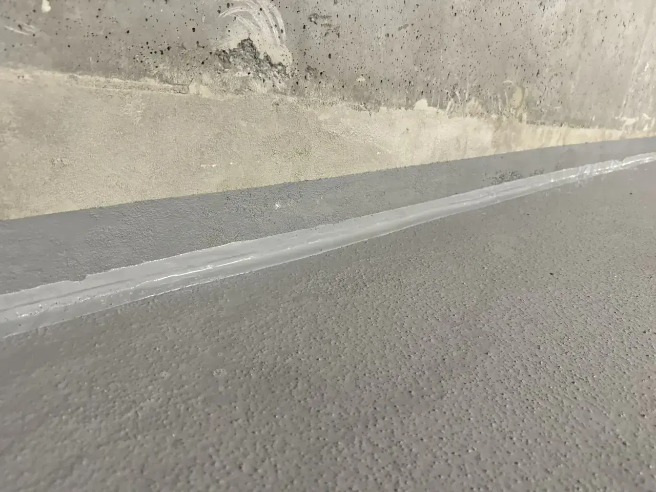 Szara posadzka z widoczną dylatacją wypełnioną białym materiałem, granicząca z betonową ścianą.