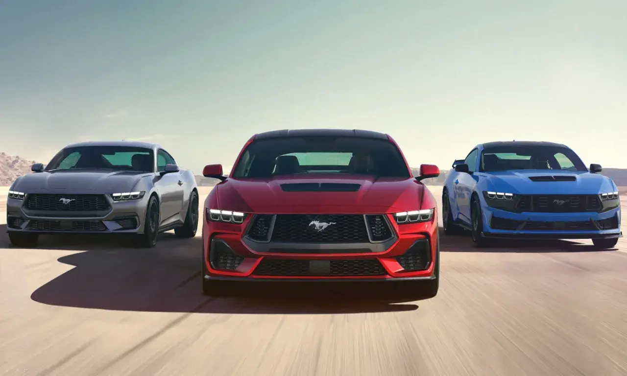 Tre Ford Mustang modeller i rött, grått och blått kör på en ökenväg.