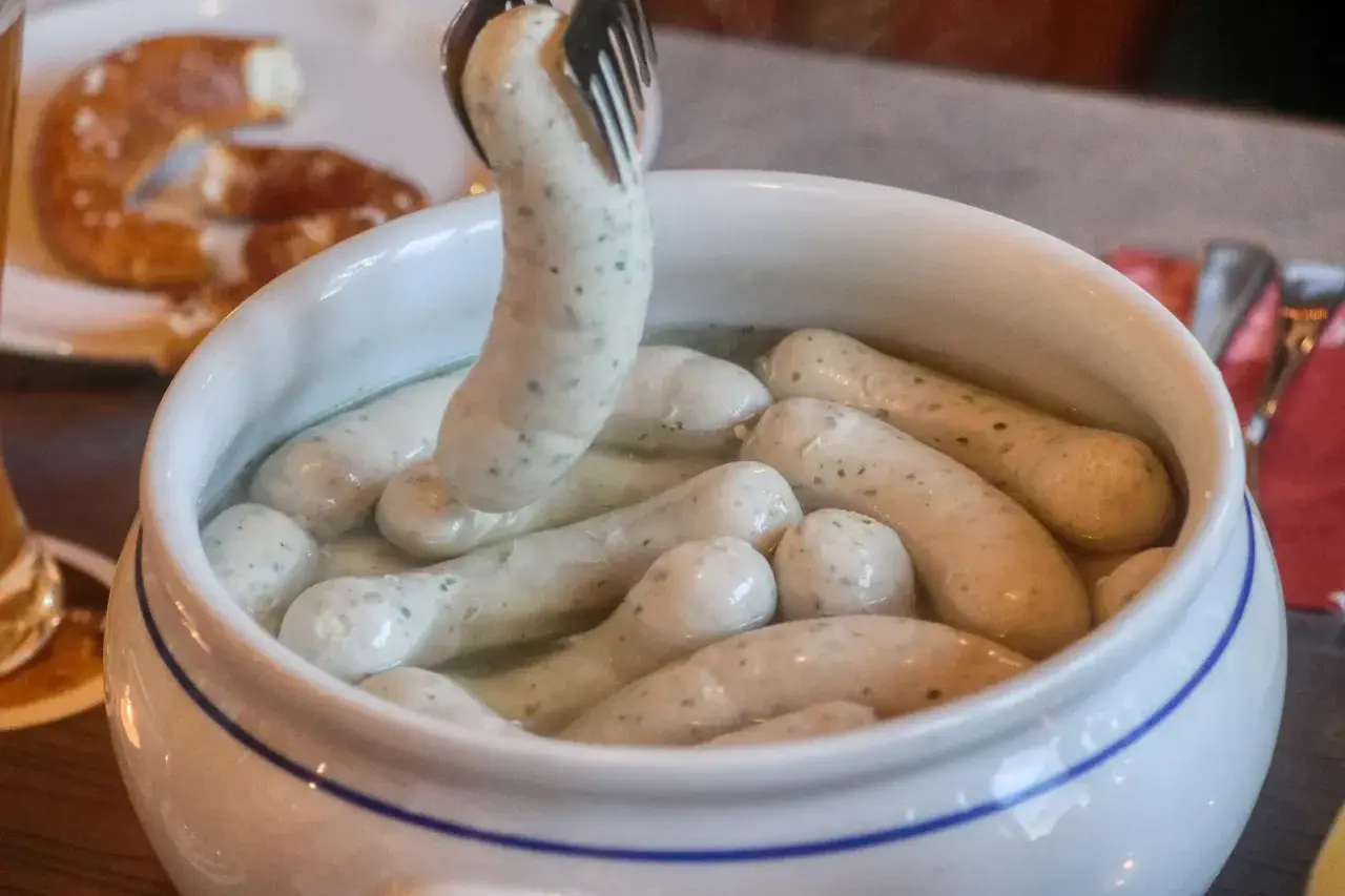 Eine Gabel hebt eine Weißwurst aus einem Topf, um sie zuzubereiten. Daneben liegt eine Brezel.
