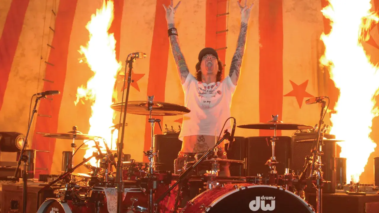 Tommy Lee w ogniu, z rękami w górze i pałeczkami perkusyjnymi, na tle płonącej sceny.