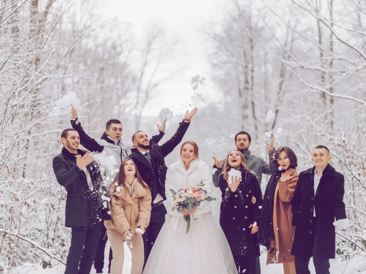Brautpaar und Gäste werfen Schnee in der Luft. Perfekt für eine Hochzeit im Winter: Was anziehen? Warme Mäntel und festliche Kleidung.