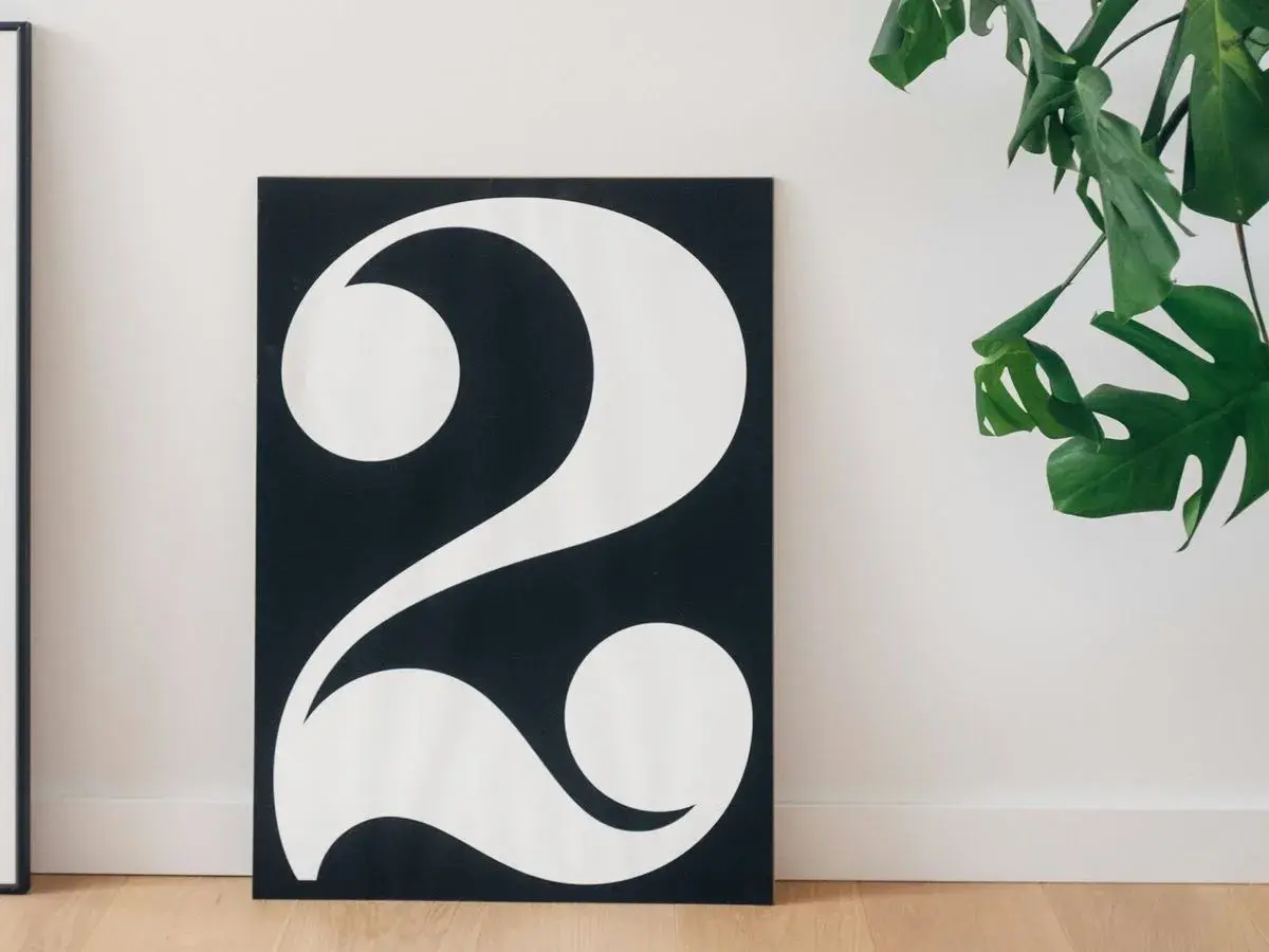 Póster minimalista con el número 2 en blanco sobre fondo negro, evocando su 2 significado espiritual. Una planta verde decora la escena.