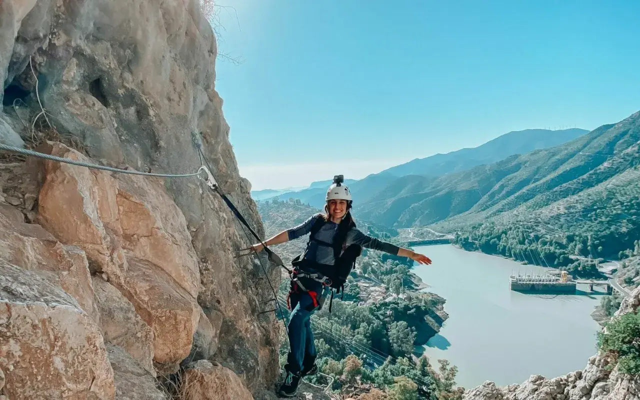 Aventura en el Caminito del Rey. Mujer sonríe mientras recorre la vía ferrata, con un embalse y montañas de fondo.