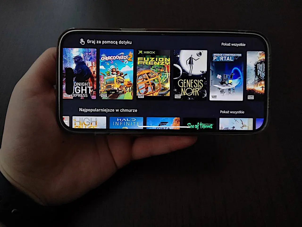 Ręka trzyma smartfon z grami do samolotu. Widoczne okładki gier: DUSK, Overcooked! 2, Fuzion Frenzy, Genesis Noir, Bridge Constructor Portal, High On Life, Halo Infinite, Forza Horizon, Sea of Thieves.