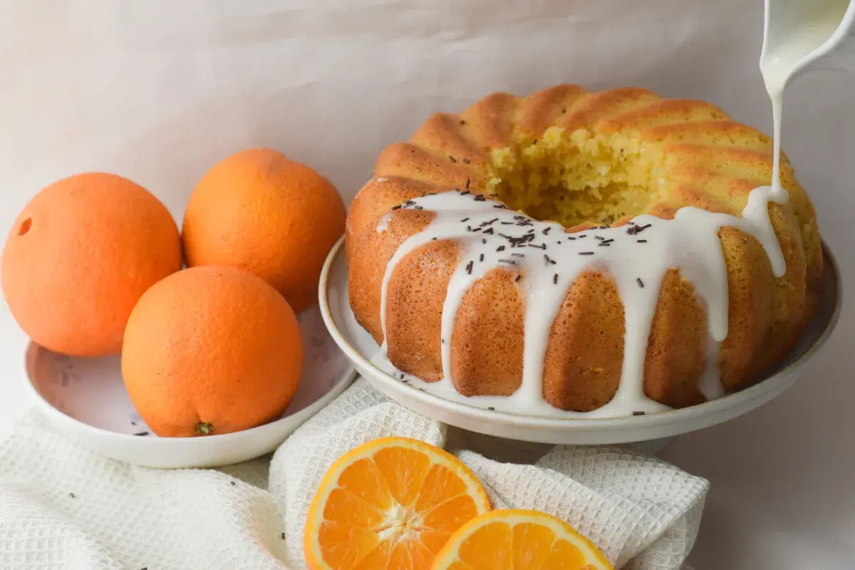 Bizcocho de naranja y yogur con glaseado blanco y chispas de chocolate. Naranjas frescas y rodajas de naranja completan la escena.