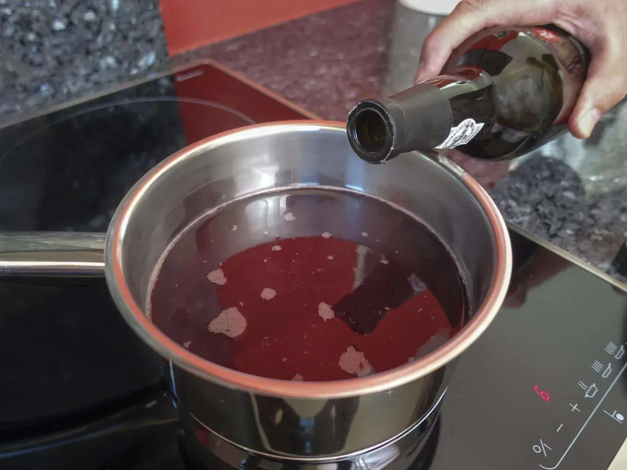 Eine Hand gießt Rotwein in einen Topf, um ihn zu kochen. Der Herd zeigt die Einstellung "LO".