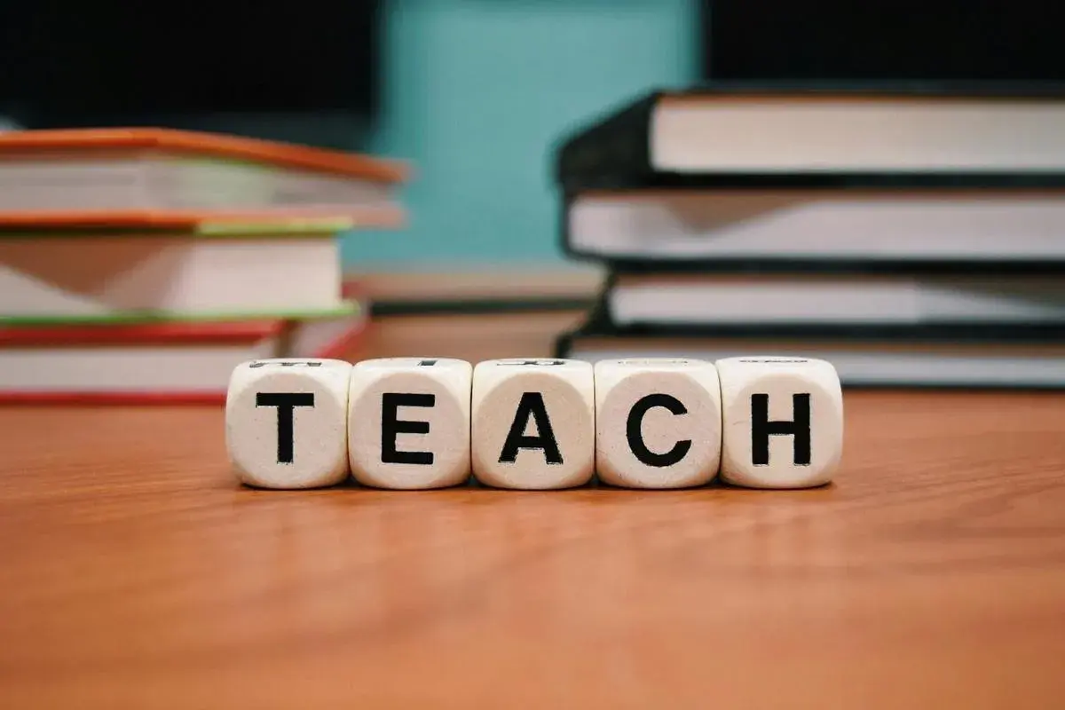 Klocki z literami "TEACH" symbolizują naukę i kolejność wprowadzania liter w klasie 1. W tle stosy książek.