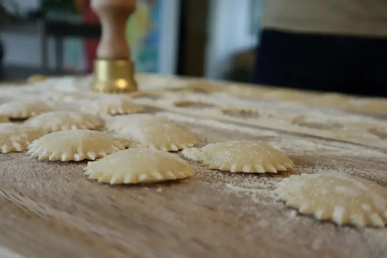 Świeżo przygotowane domowe ravioli na drewnianym blacie, posypane mąką. W tle widoczny przyrząd do wycinania pierogów.