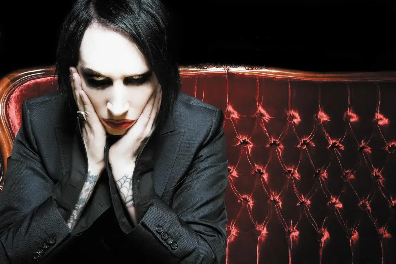Marilyn Manson, z charakterystycznym makijażem, siedzi na aksamitnej kanapie. Jego utwory często poruszają mroczne tematy.
