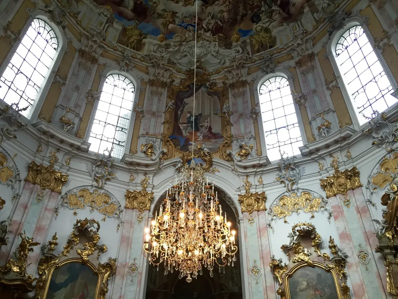Prunkvoller Kronleuchter unter einer Decke mit Fresken, umgeben von opulenten Verzierungen, die die **Rokoko Architektur** widerspiegeln.