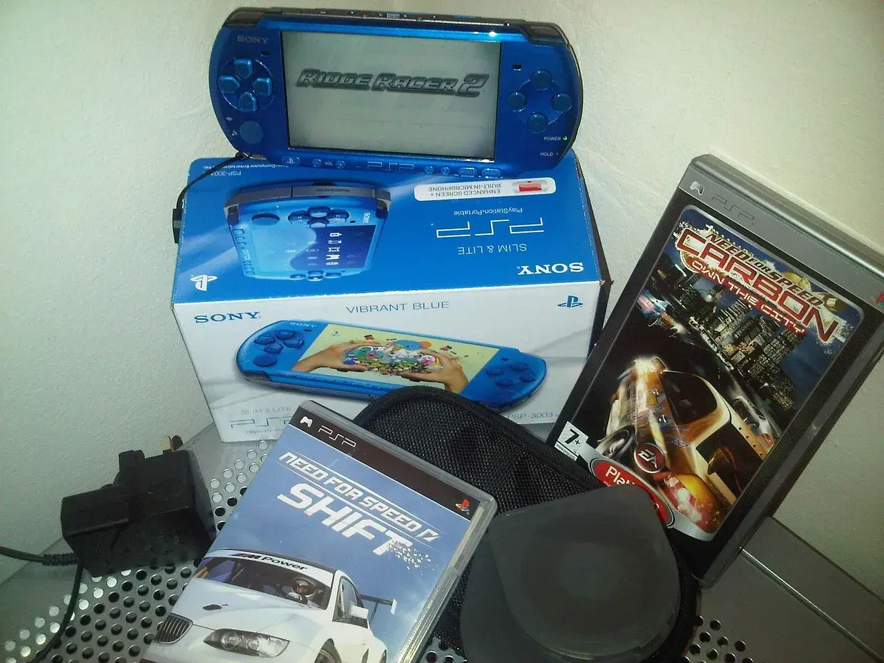 Niebieska konsola PSP z grą Ridge Racer 2, pudełko z konsolą i grami Need for Speed Shift i Carbon. Klasyczne stare gry na telefon.