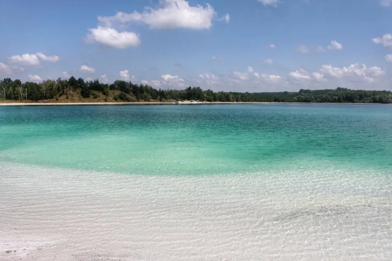 Lazurowe jezioro z piaszczystą plażą i lasem w tle. Woda jest krystalicznie czysta, a dno widoczne.