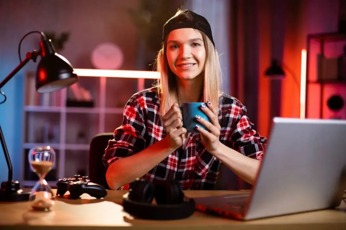 Uśmiechnięta dziewczyna z kubkiem w dłoni, przed nią laptop i kontroler do gry. Może szuka kodu na cas w The Sims 4?