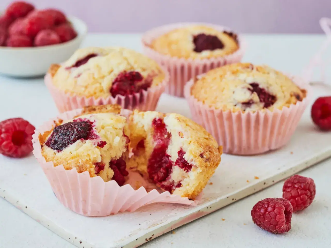Saftige Himbeer Muffins in rosa Förmchen, einer aufgeschnitten, mit frischen Himbeeren garniert.