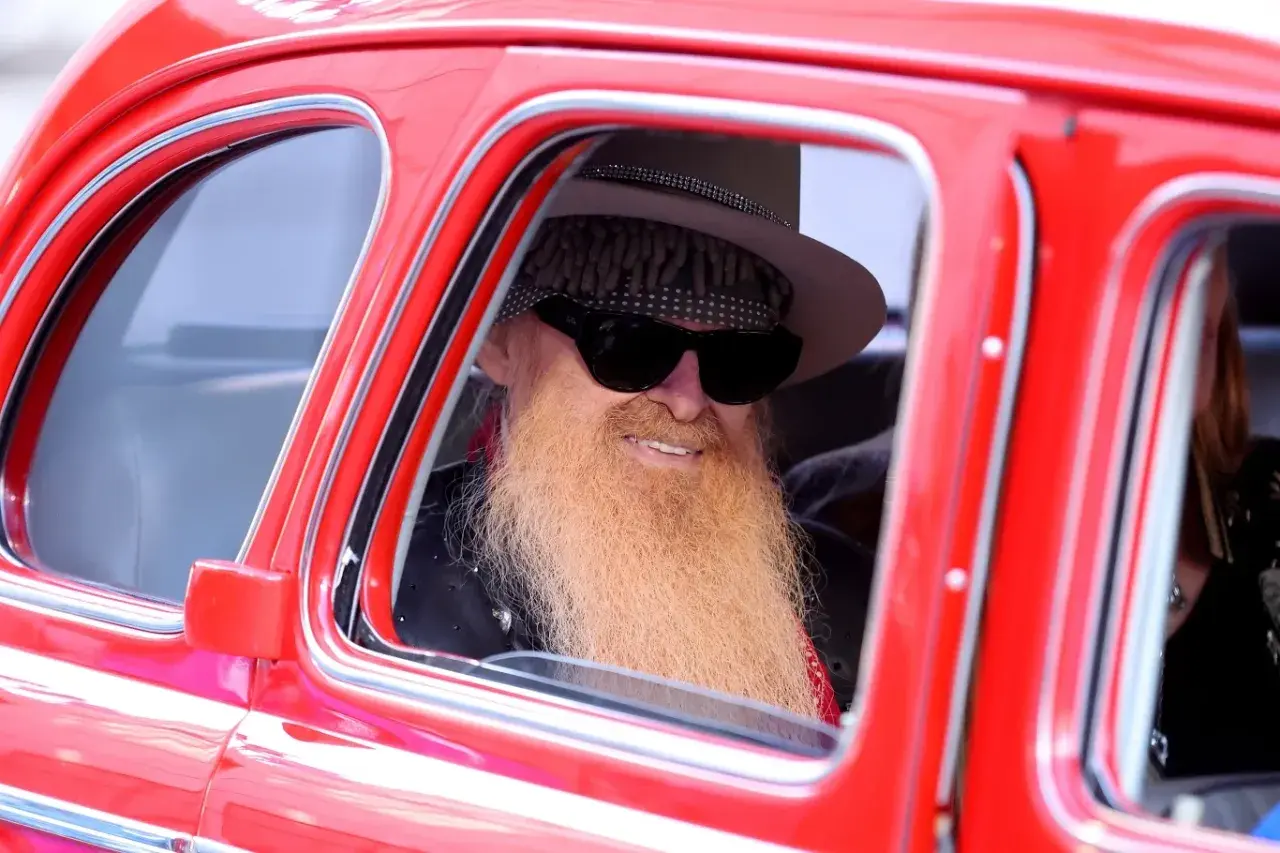 Billy Gibbons, jeden z członków ZZ Top, w stylowym kapeluszu i z charakterystyczną brodą, siedzi w czerwonym samochodzie.