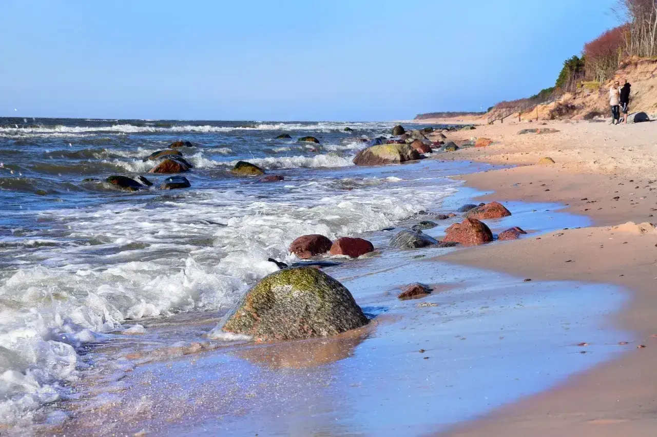 Poddąbie plaża. Fale rozbijają się o kamienie na piaszczystym brzegu. W oddali widać spacerującą parę.