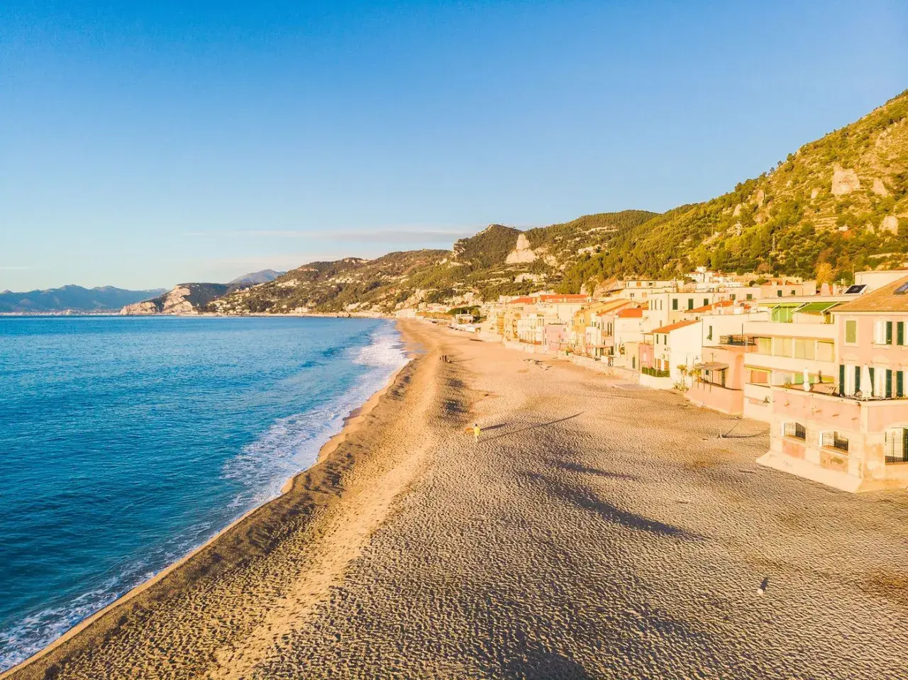 Plage de sable fin bordée de maisons colorées sur la côte italienne. La mer bleue et les collines verdoyantes complètent ce paysage paisible.
