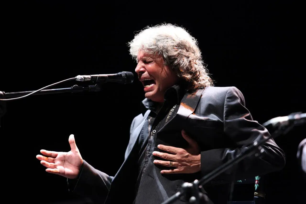Un cantante de grupos flamencos de los 80, con melena rubia, canta apasionadamente frente a un micrófono, con la mano en el pecho.