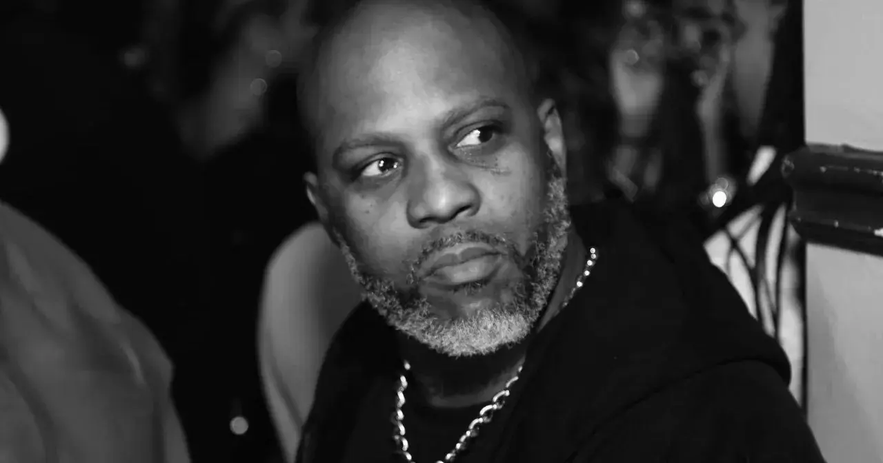 DMX, z siwą brodą i łańcuchem na szyi, spogląda w bok. W tle widać niewyraźne sylwetki, być może jego dzieci.