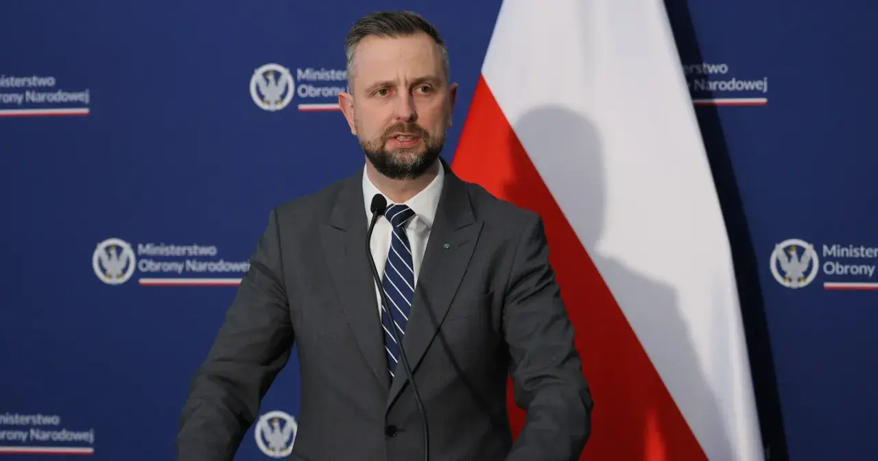 Mężczyzna w garniturze, minister obrony narodowej, przemawia przy mikrofonie na tle flagi Polski i logo ministerstwa.