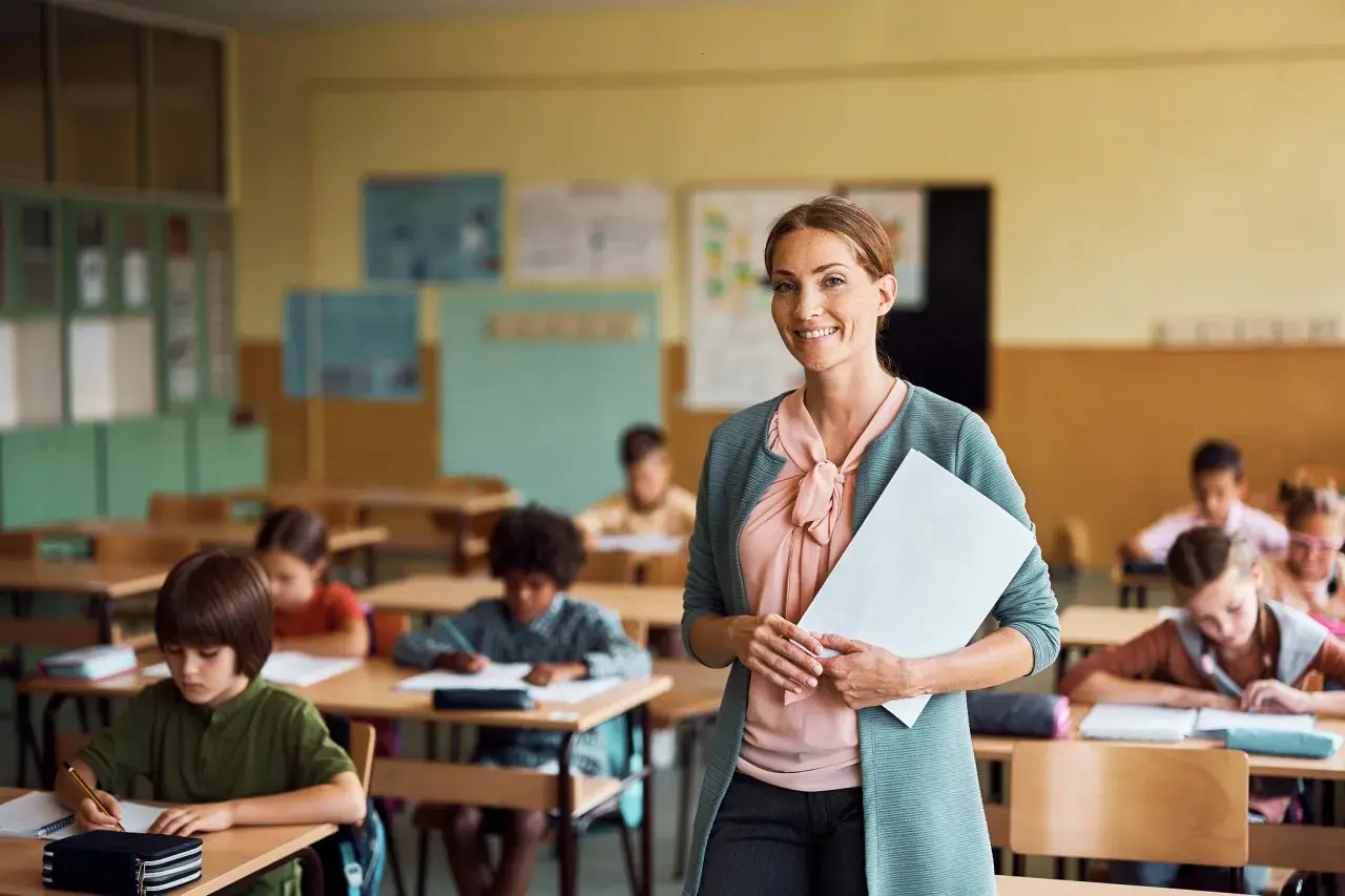 Lächelnde Lehrerin mit Unterlagen in einem Klassenzimmer voller konzentrierter Kinder. Sie fragt sich: "Soll ich Lehrer werden?