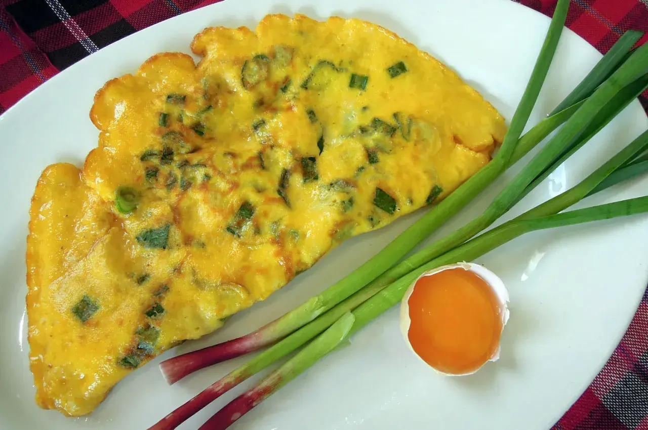 Une omelette à l'aillet, garnie de ciboule verte, est servie sur une assiette blanche avec des tiges d'aillet et un jaune d'œuf.