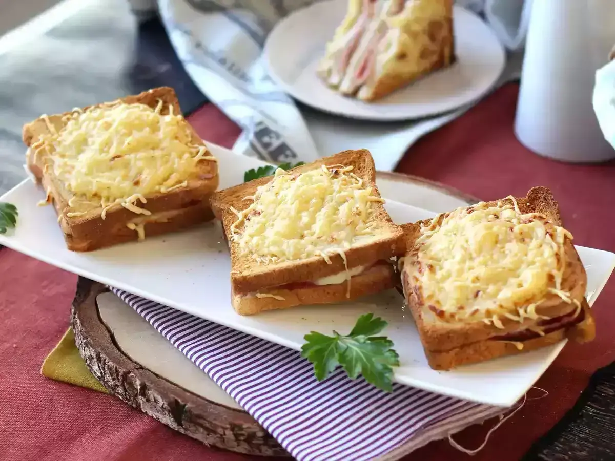 Tre gyllene croque monsieur, gratinerade med ost, ligger uppradade på en vit bricka. En klassisk fransk delikatess.