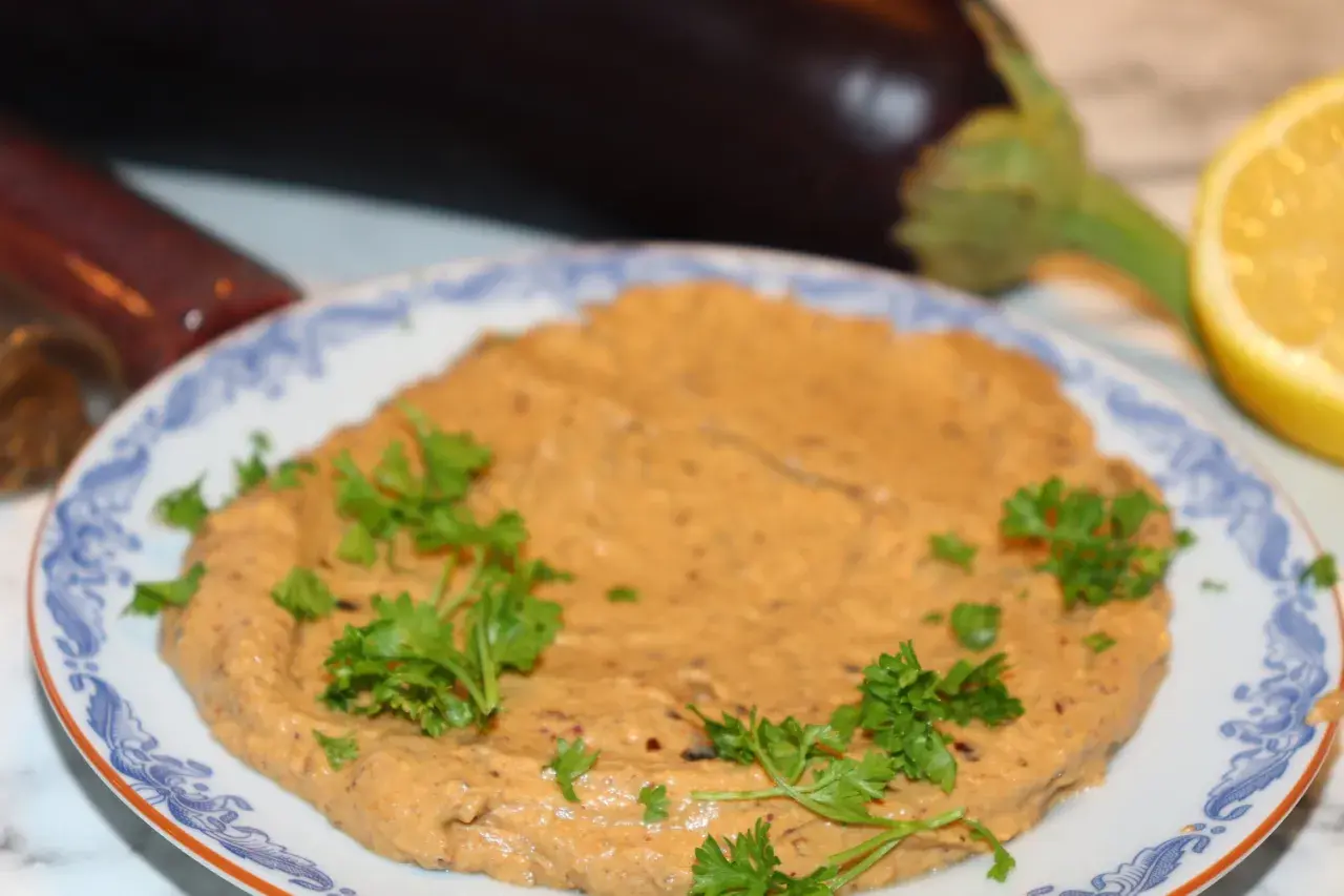 En krämig baba ganoush, garnerad med persilja, med aubergine och citron i bakgrunden.