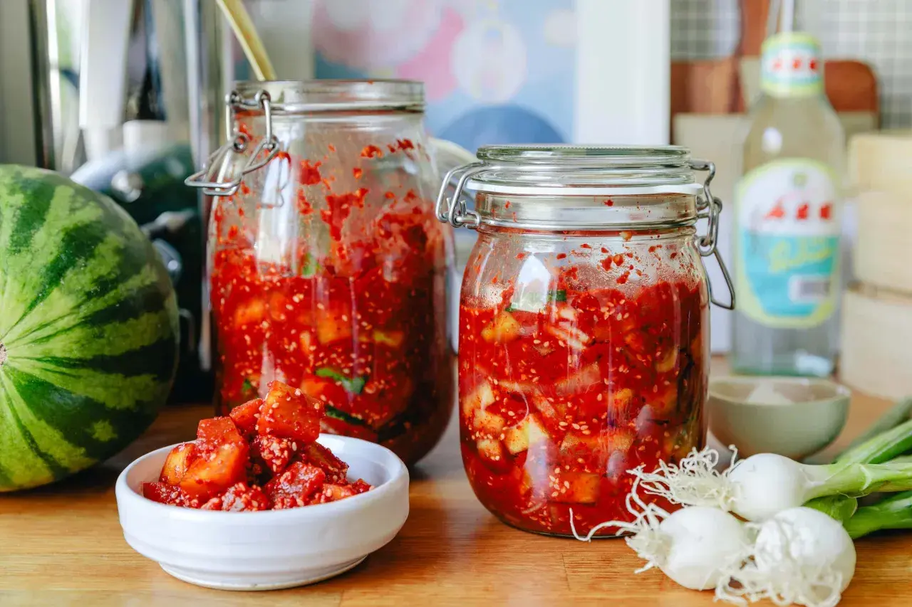 Zwei Gläser mit fermentierter Melone, daneben eine Wassermelone, Frühlingszwiebeln und eine Flasche.