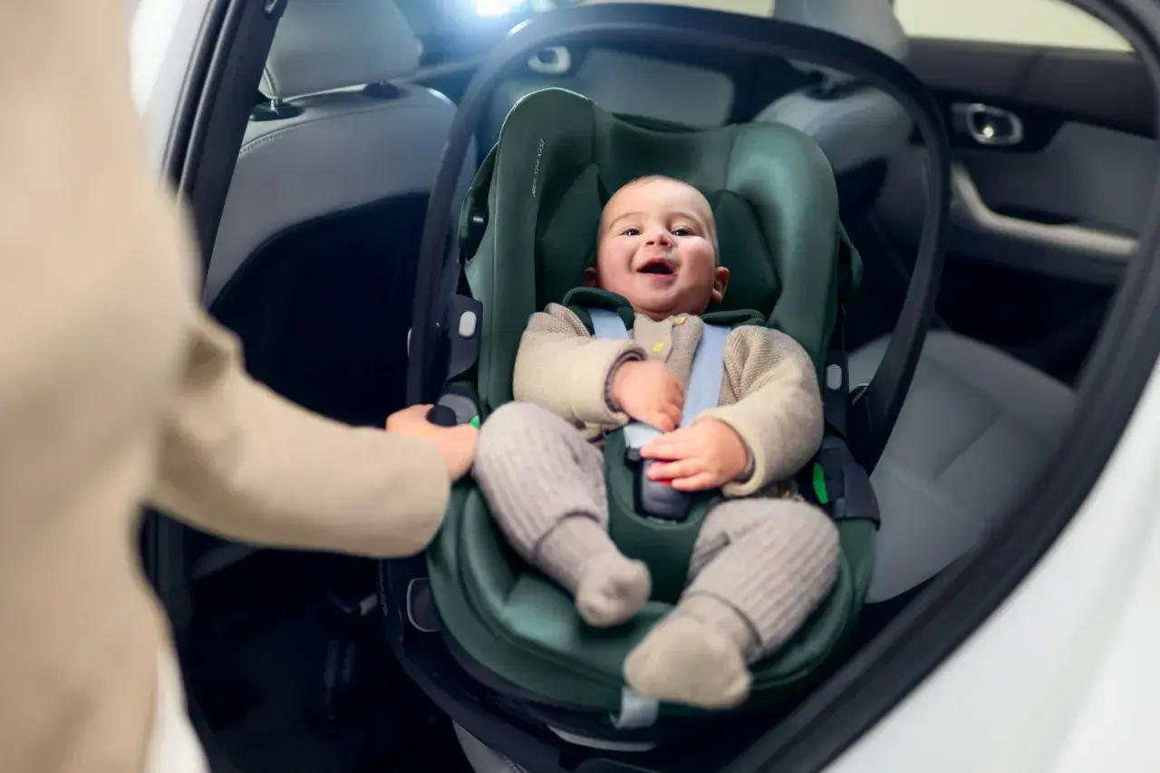 Uśmiechnięte dziecko w foteliku samochodowym. Ręka rodzica pokazuje, jak wypiąć fotelik z bazy isofix.