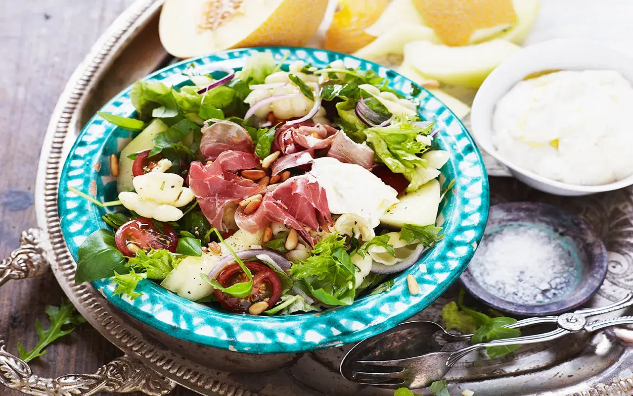 En härlig ost och skinksallad med melon, tomater och pinjenötter. Serveras på en vacker turkosblå skål.