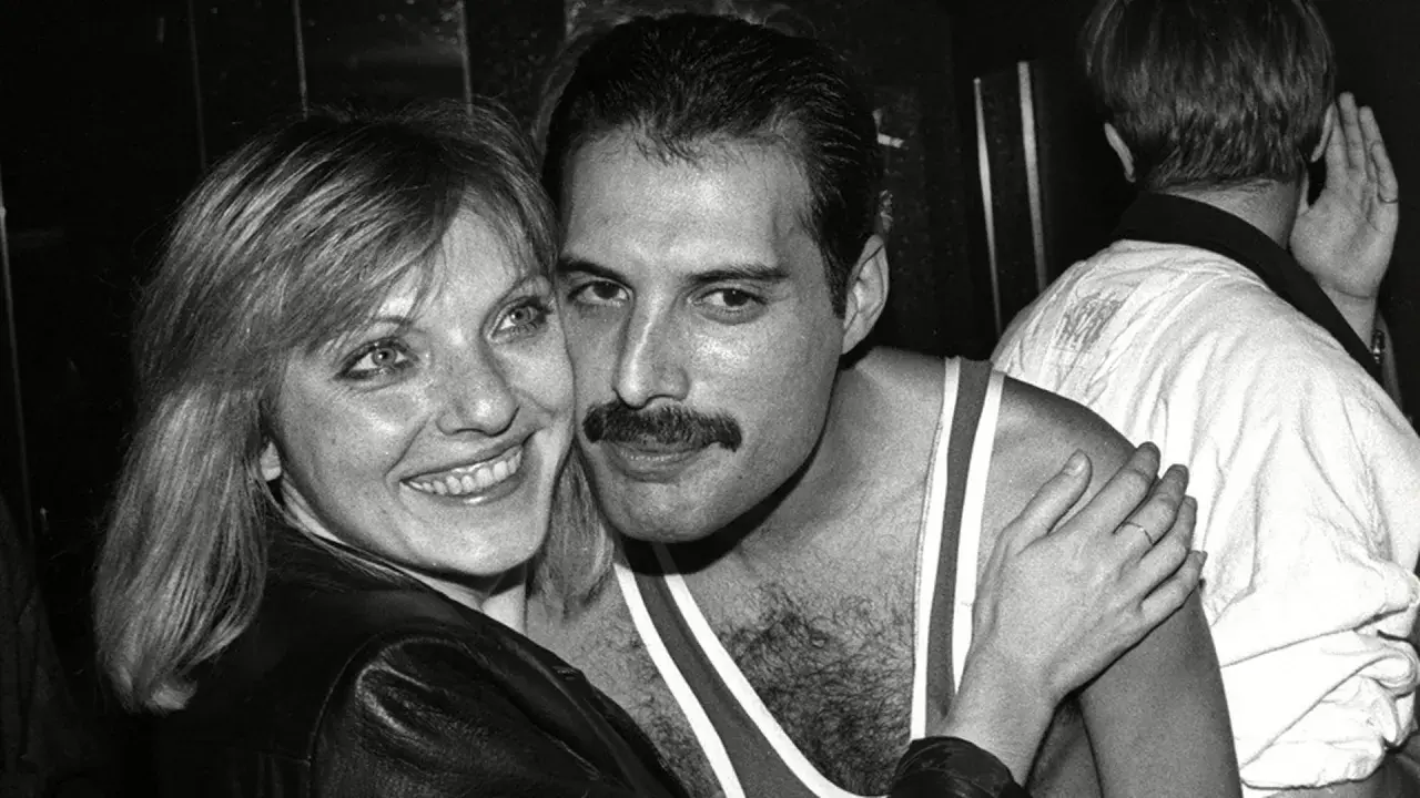 Freddie Mercury z uśmiechem, odsłaniającym jego charakterystyczne zęby, przytula się do kobiety.