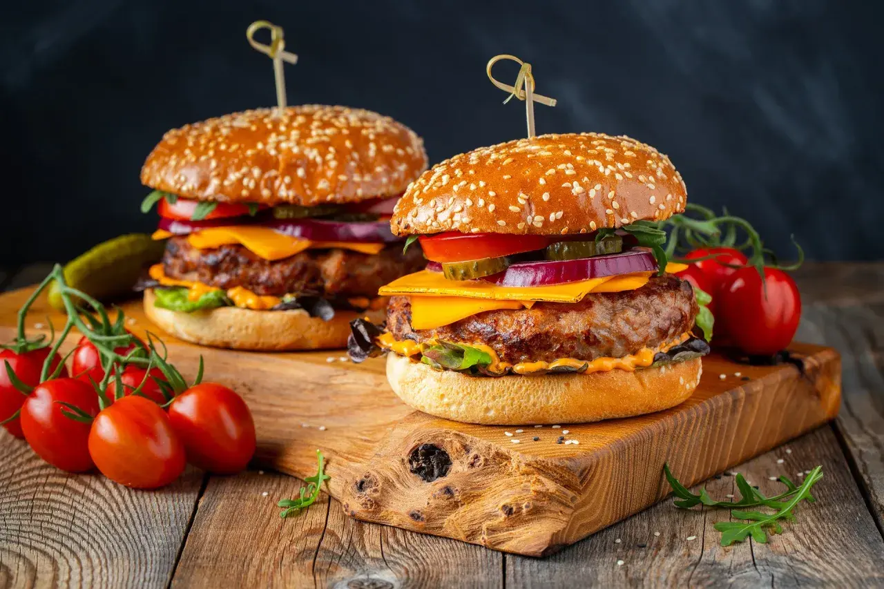 Dwa soczyste burgery z idealnie wysmażonym mięsem na burgery, serem, warzywami i sezamową bułką, na drewnianej desce.