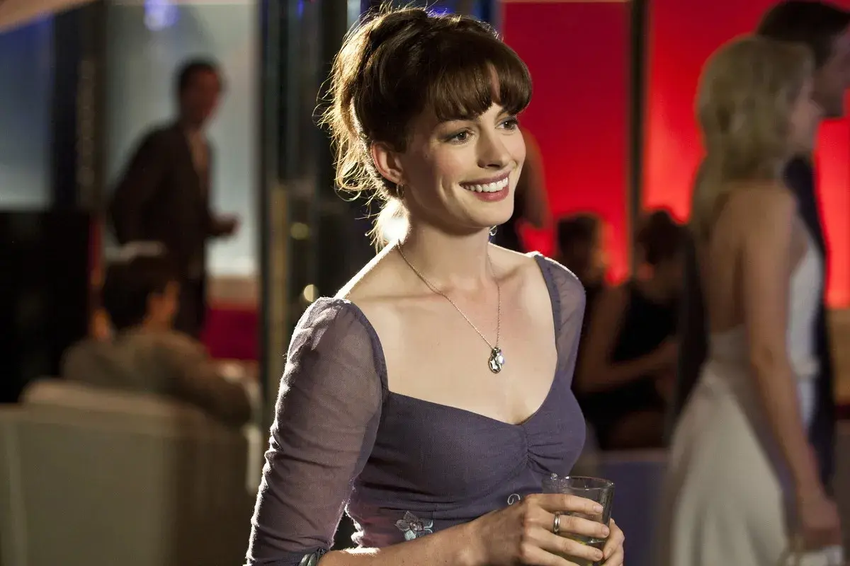 Anne Hathaway w filmie, uśmiechnięta, trzymająca drinka.