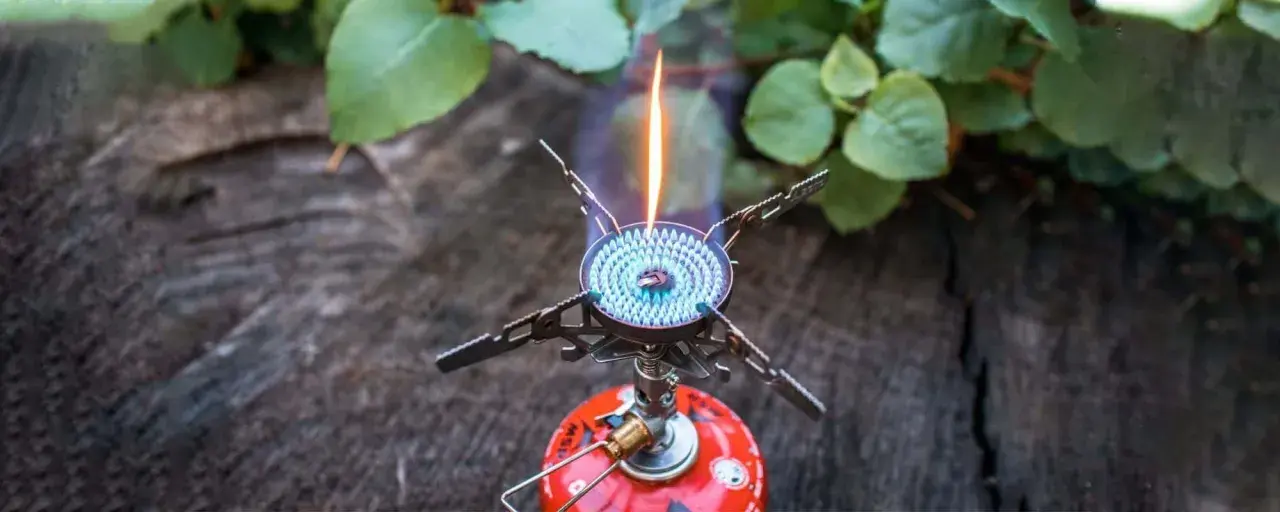 Soto Windmaster Kocher mit blauer Flamme auf einem Baumstamm, umgeben von grünen Blättern.