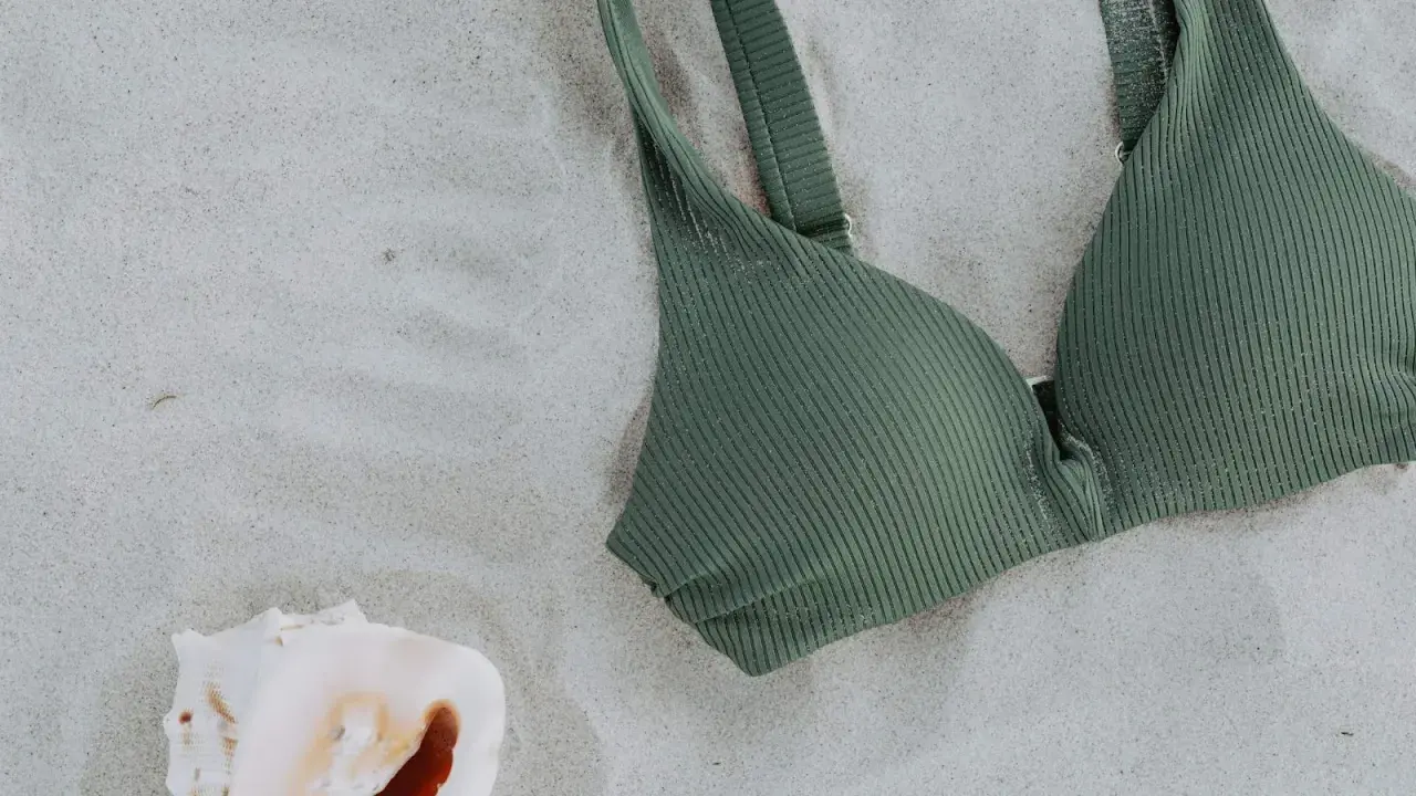A ribbed green swim fabric bikini top rests on the sand next to a seashell.