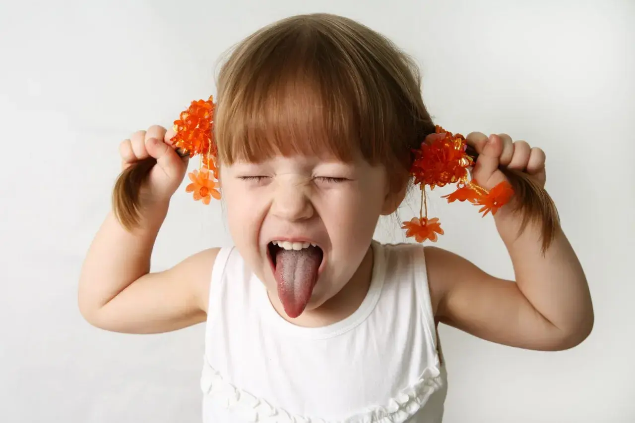 Bambina iperattiva fa una smorfia con la lingua fuori, tirando le trecce con fiori arancioni. Cosa fare?