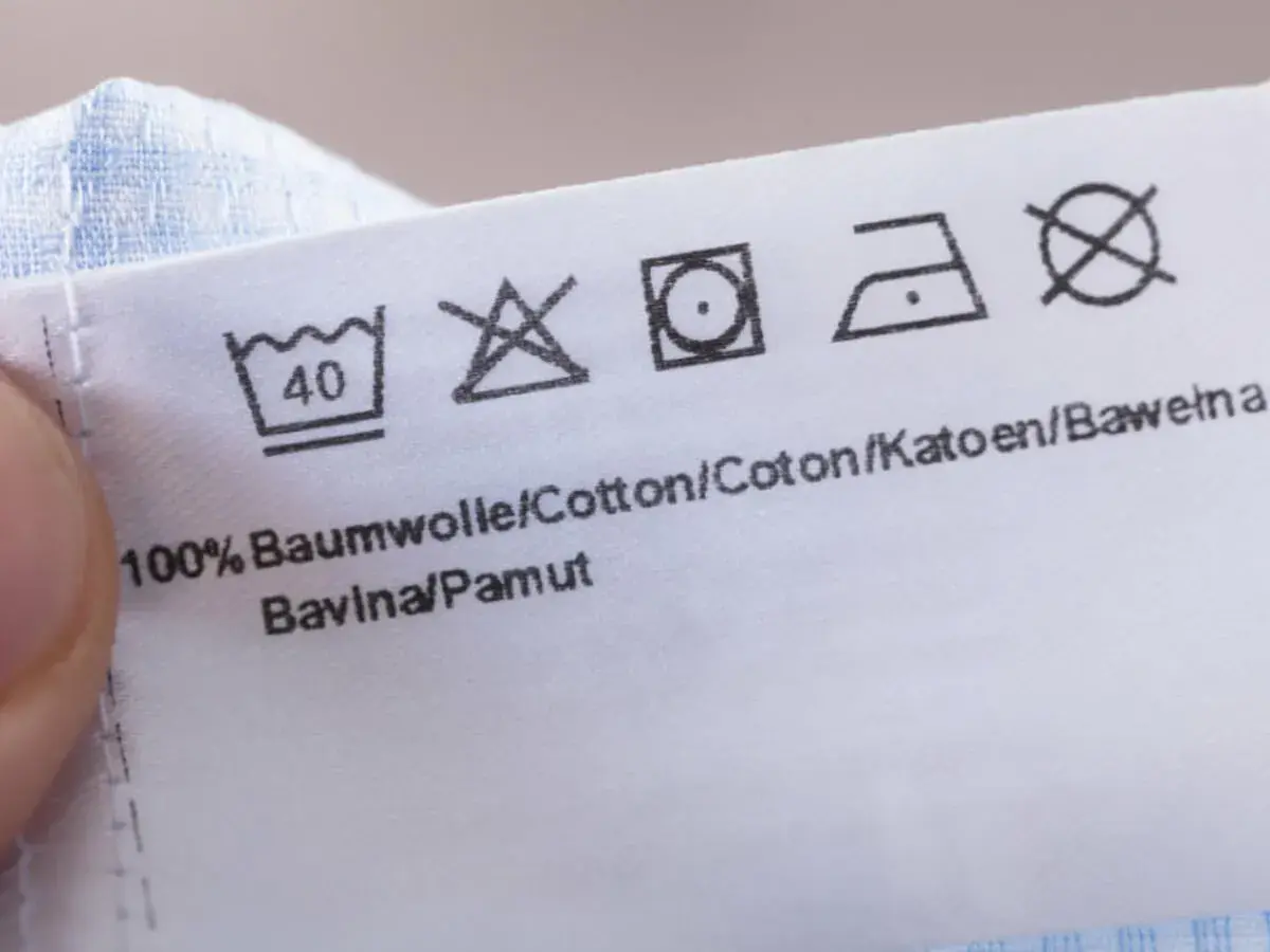 Pflegeetikett mit Waschsymbolen und dem Hinweis "100% Baumwolle". Die Symbole zeigen, dass das Kleidungsstück bei 40°C waschbar ist, nicht bleichen, bei niedriger Temperatur im Trockner trocknen und bei mittlerer Hitze bügeln.
