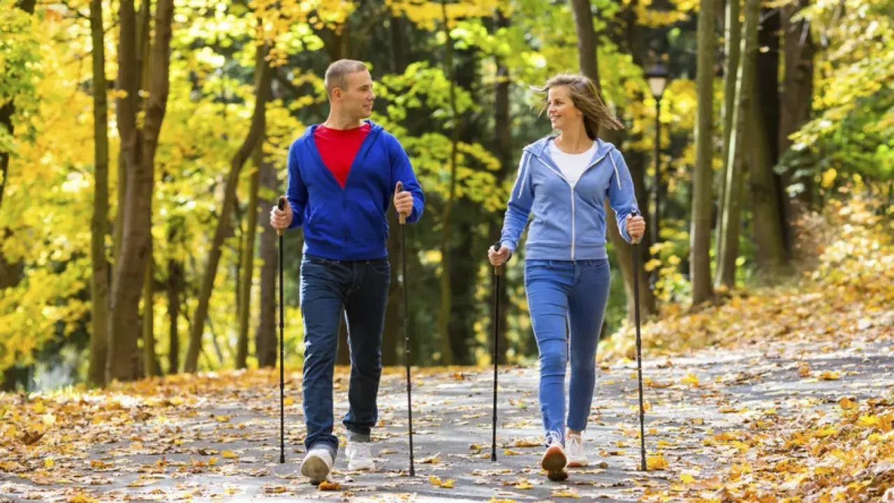 Para spaceruje po jesiennym parku, uprawiając nordic walking. Liście tworzą kolorowy dywan.