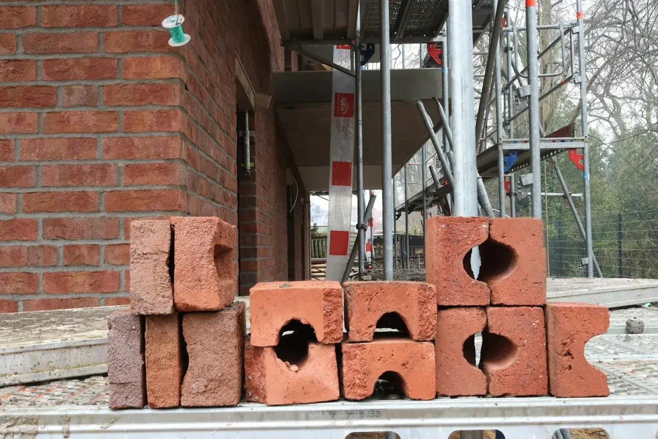 Stos cegieł na budowie. W tle widać fragment czerwonej ściany i rusztowania.