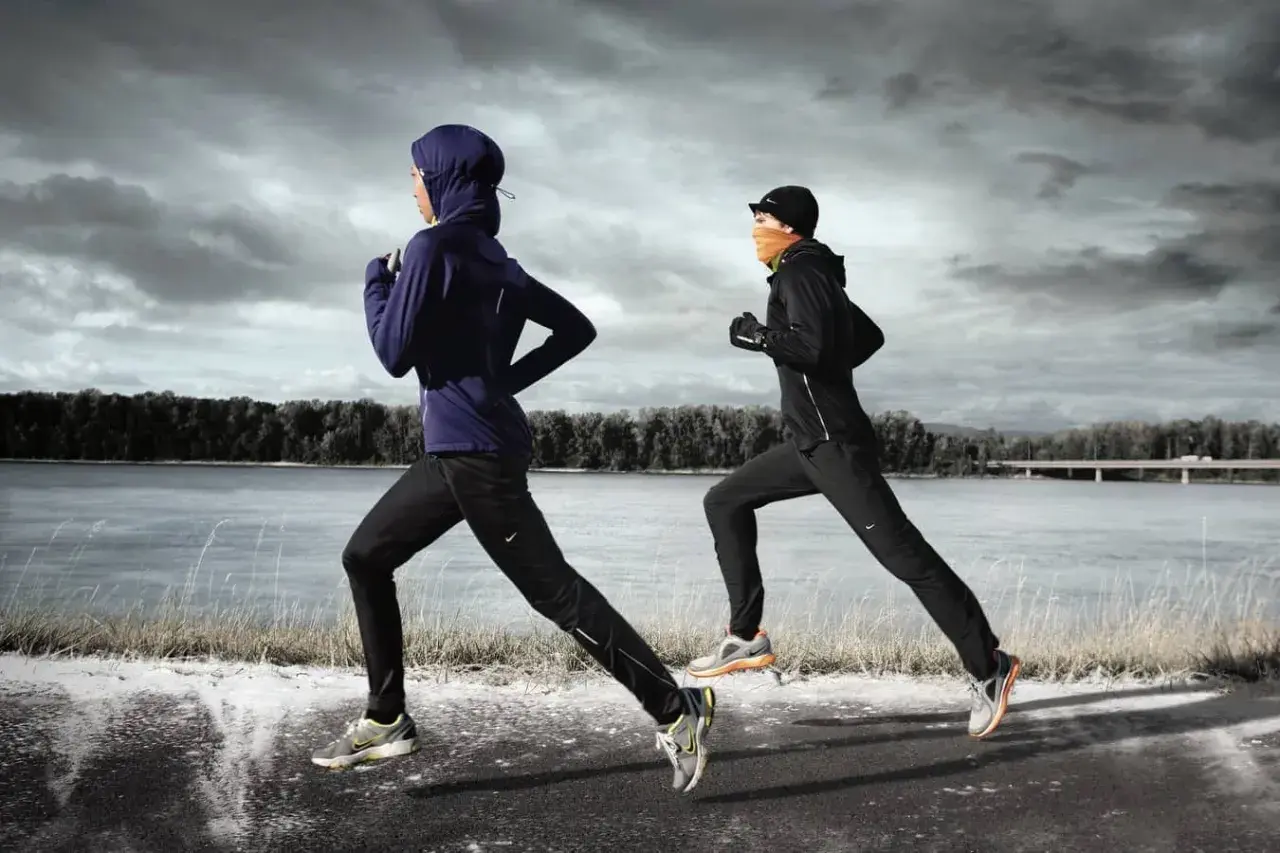 Zwei Läuferinnen in warmer Winterkleidung joggen am Fluss entlang. Die eine trägt einen blauen Hoodie und Hijab, die andere eine schwarze Jacke und orangefarbenen Schal.