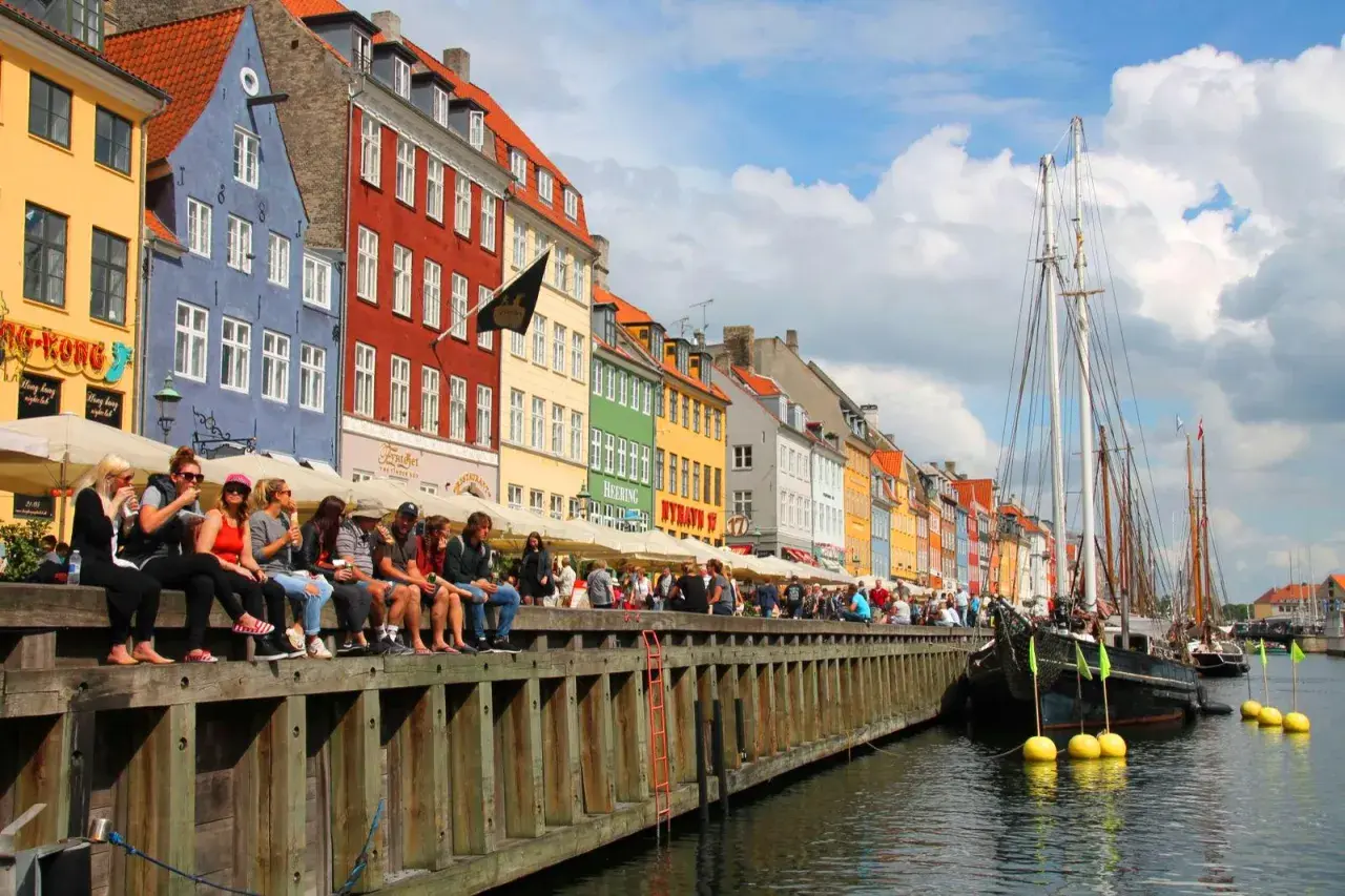 Bunte Häuser am Nyhavn, Kopenhagen an einem Tag. Menschen sitzen am Kai, Boote liegen im Wasser.