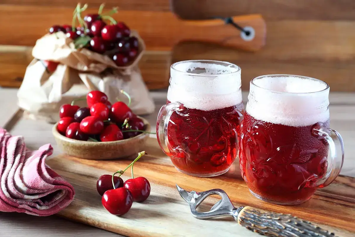 Due boccali di birra kriek rossastra con schiuma bianca, circondati da ciliegie fresche su un tagliere di legno.