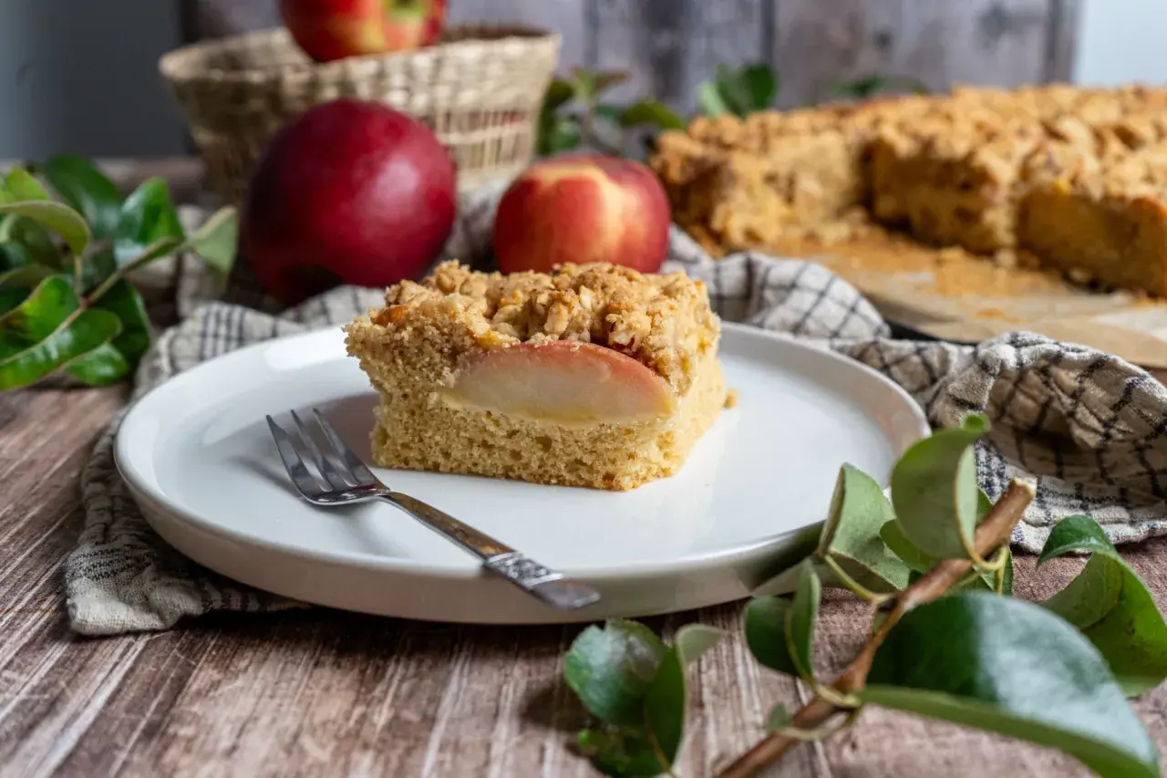 Ein Stück saftiger Apfelkuchen mit Streuseln, perfekt für ein Apfelringe Rezept. Daneben liegen frische Äpfel.