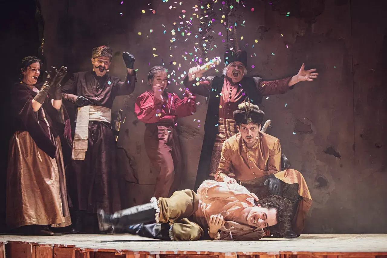 Aktorzy w strojach ludowych świętują na scenie, sypiąc konfetti. To fragment repertuaru teatru ludowego.