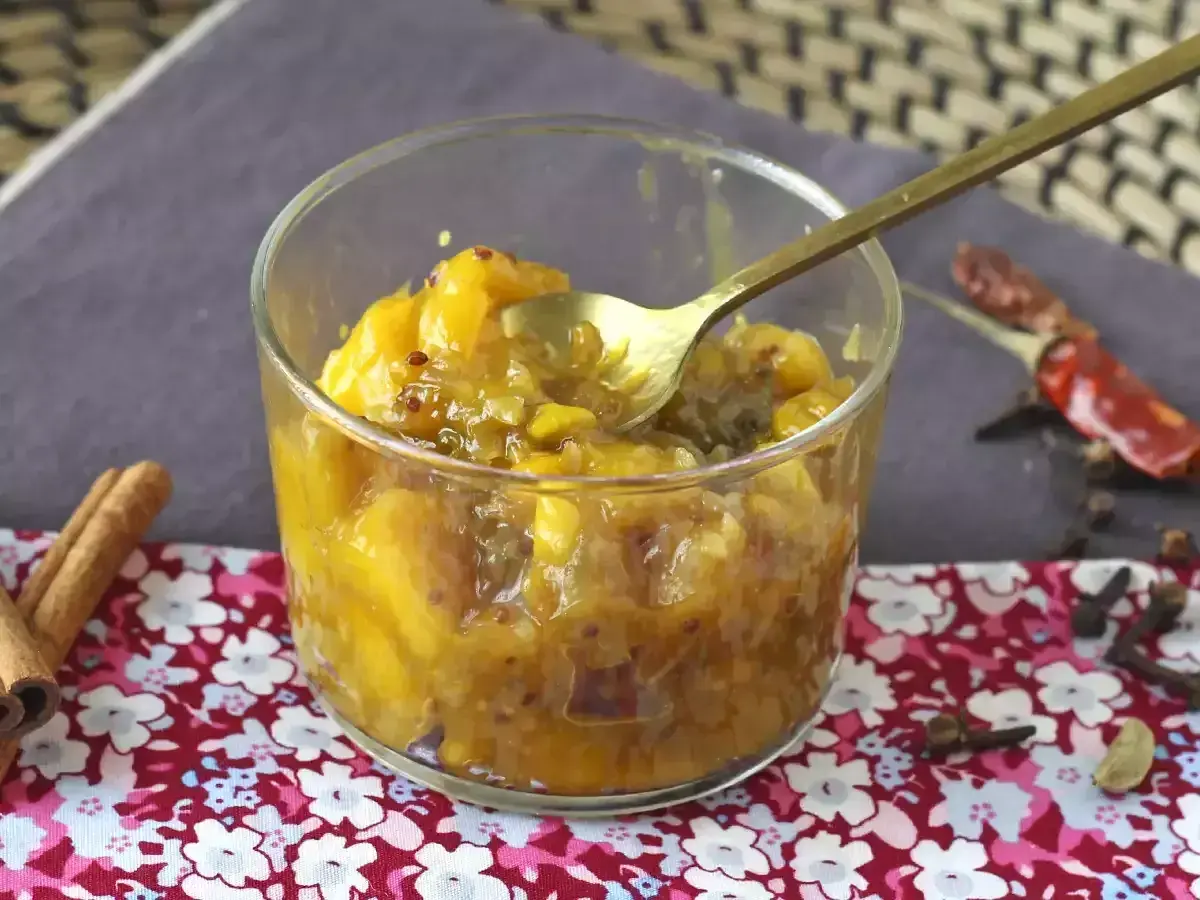Un verre rempli de chutney de mangue, avec une cuillère dorée. Des épices comme la cannelle, le piment et le clou de girofle entourent le plat.