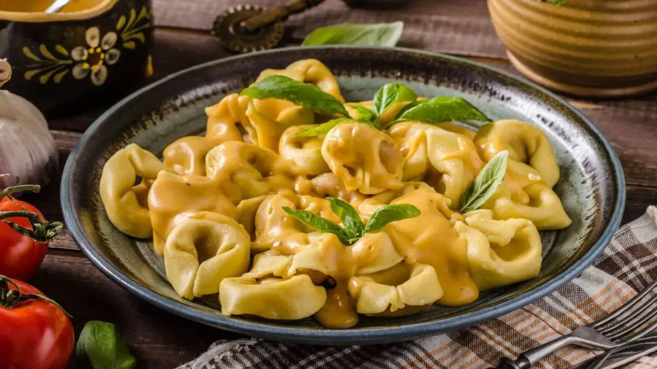 Plato de tortellini de carne cubiertos con una cremosa salsa de queso y decorados con hojas de albahaca fresca.