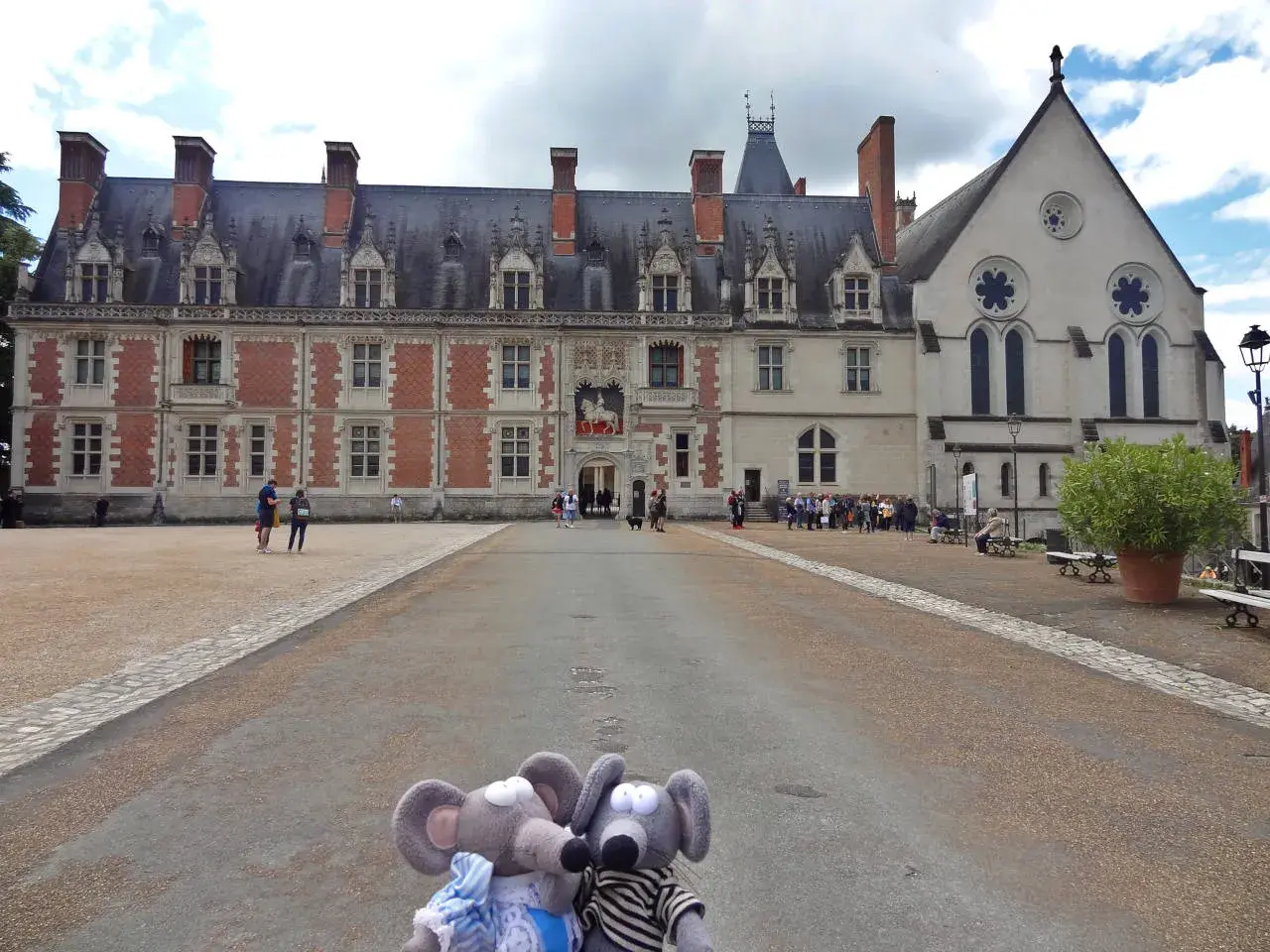 Deux souris en peluche devant le majestueux **château de Blois**. Des visiteurs explorent la cour de ce monument historique.
