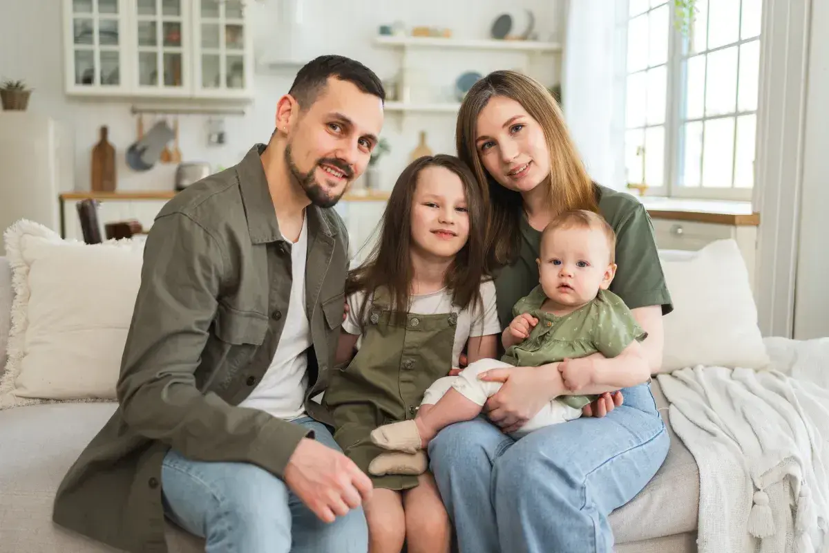 Szczęśliwa rodzina z dwójką dzieci siedzi na kanapie. Rodzice i dzieci uśmiechają się. Program 800 plus obejmuje dzieci do 18 roku życia.