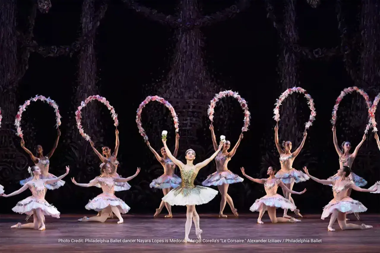 Balettdansöser i ljusa dräkter utför en scen från "Le Corsaire", med blomsterkransar i händerna.